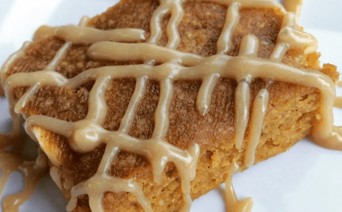 A close-up of a square piece of dessert drizzled with a creamy glaze on a white plate.