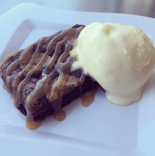 A rich chocolate brownie drizzled with caramel sauce, served with a scoop of vanilla ice cream on a white plate.