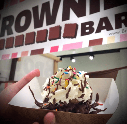 A hand holds a dessert topped with whipped cream, chocolate drizzle, and sprinkles, in front of a "Brownie Bar" sign.