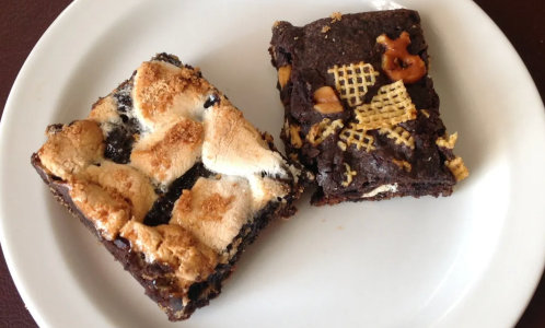 Two chocolate dessert bars on a white plate, one topped with marshmallows and the other with a crunchy texture.