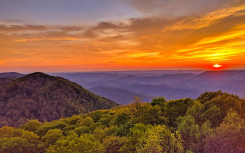 A vibrant sunset over rolling mountains, with layers of trees and hills silhouetted against a colorful sky.