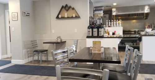 A cozy café interior with metal chairs, wooden tables, and a counter featuring coffee machines and a grill in the background.