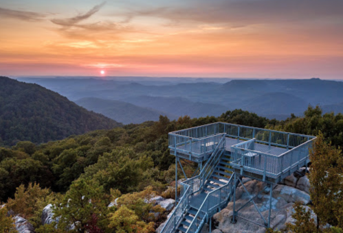 A viewing platform on a mountain at sunset, overlooking rolling hills and a colorful sky.