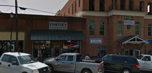 A street view of a brick building featuring "Fortify Kitchen & Bar" and other shops, with parked trucks in front.