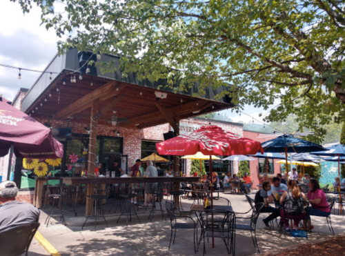 Outdoor dining area of a restaurant with umbrellas, tables, and people enjoying their meals under a tree.