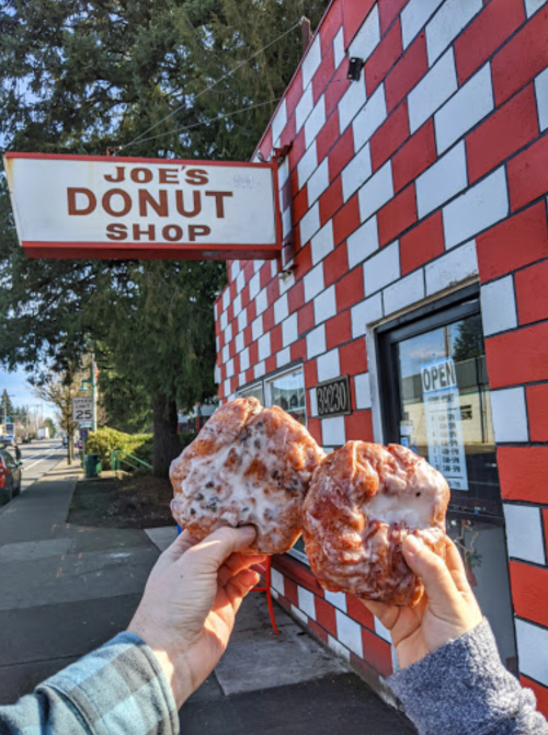 Two hands hold glazed donuts in front of Joe's Donut Shop, featuring a red and white checkered exterior.