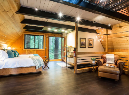 Cozy wooden cabin interior with a bed, armchair, and large windows overlooking trees, featuring warm lighting.