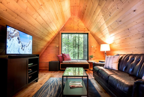 Cozy cabin interior with wooden walls, a black leather sofa, a coffee table, and a large window overlooking trees.