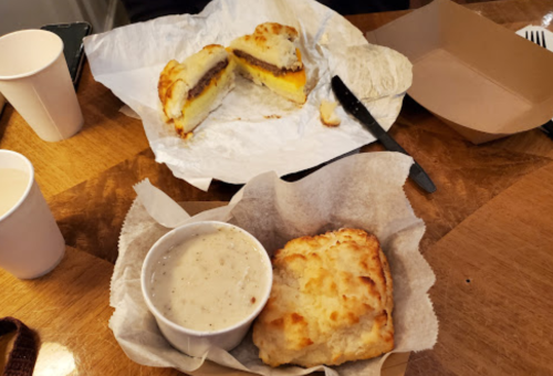 A plate with a biscuit sandwich filled with sausage and cheese, alongside a bowl of creamy soup and a paper cup.