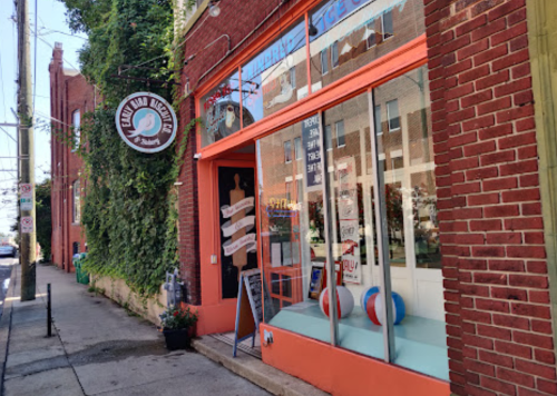 A colorful storefront with a sign, large windows, and beach balls outside, surrounded by greenery on a sunny day.