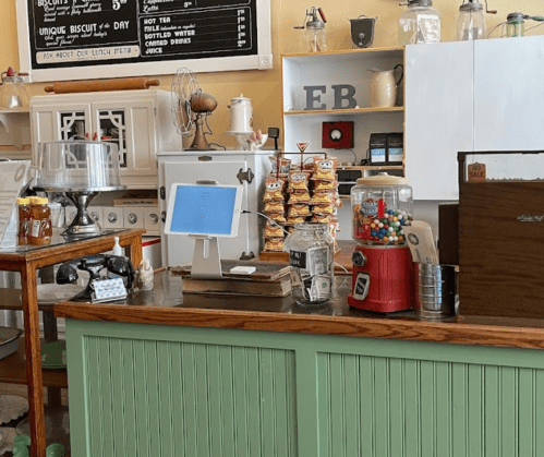 A cozy café counter with a cash register, snacks, a gumball machine, and a menu board in a warm, inviting setting.
