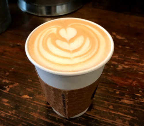 A latte in a paper cup with a heart-shaped foam design on top, resting on a wooden table.