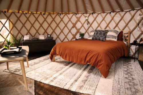 Cozy yurt interior featuring a bed with orange bedding, patterned rugs, and a wooden table with plants.