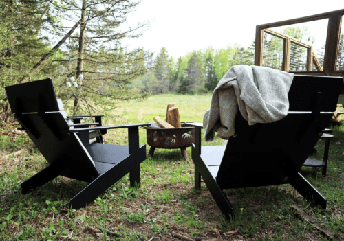 Two black outdoor chairs facing a fire pit, surrounded by greenery and trees, with a cozy blanket draped over one chair.