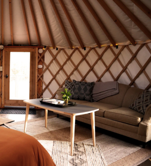 Cozy interior of a yurt featuring a sofa, coffee table, and wooden door, with warm lighting and natural decor.