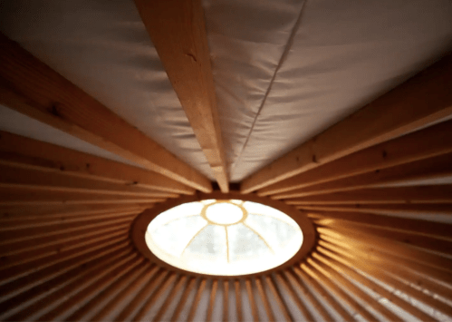 View of a circular skylight surrounded by wooden beams in a yurt, with soft light filtering through.
