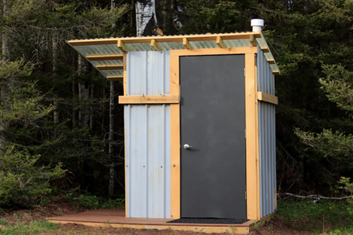 A small, rustic shed with a metal roof and wooden frame, set among trees in a natural outdoor setting.