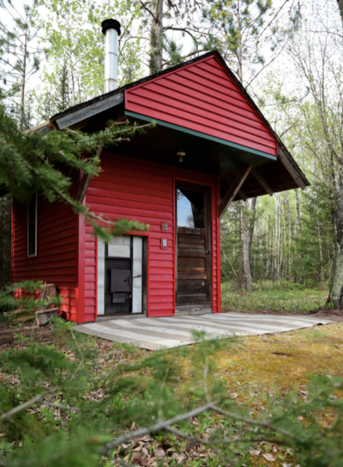 A small red cabin with a sloped roof, surrounded by trees and greenery, featuring a wooden door and a chimney.