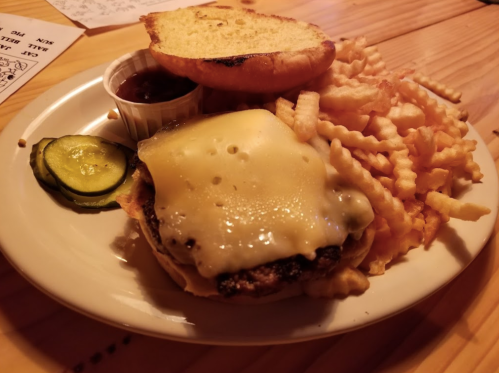 A plate with a cheeseburger topped with melted cheese, pickles, a side of crinkle fries, and a small cup of sauce.