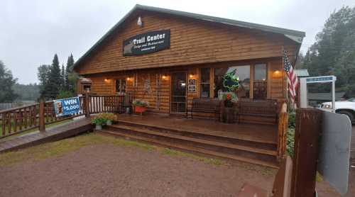 A wooden building with a sign reading "Trail Center," featuring benches, flower pots, and an American flag outside.