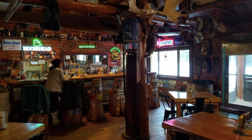 Cozy rustic bar interior with wooden decor, neon signs, and a bartender preparing drinks. Tables are set for patrons.