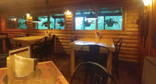 Cozy wooden restaurant interior with tables, chairs, and large windows overlooking greenery. Warm, inviting atmosphere.