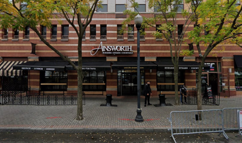 A brick building with a sign reading "Ainsworth," featuring outdoor seating and trees lining the sidewalk.
