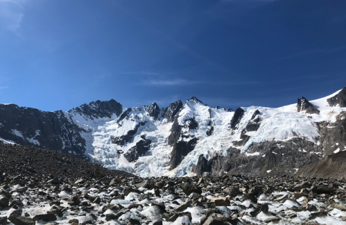 A rocky landscape with a glacier and snow-capped mountains under a clear blue sky.