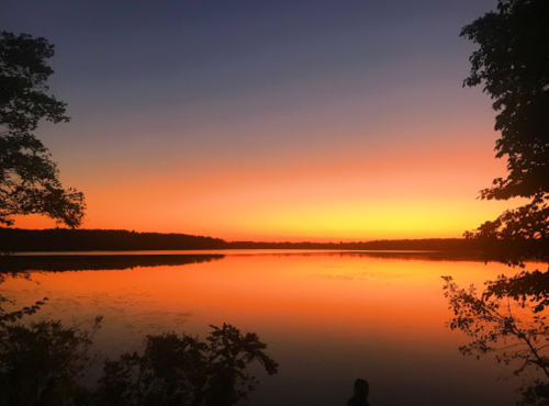 A serene lake at sunset, reflecting vibrant orange and purple hues in the sky, surrounded by trees.