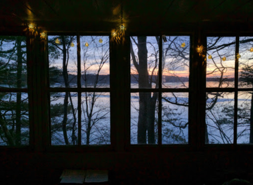 View of a serene lake at sunset through a window, framed by trees and soft glowing lights.