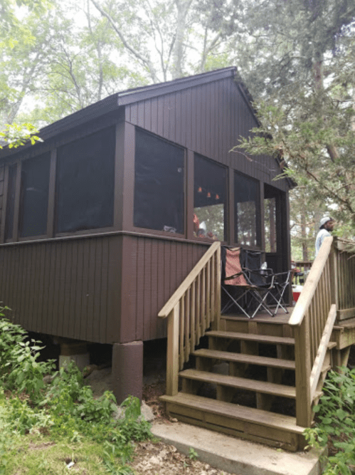A brown cabin with a screened porch, surrounded by trees, featuring a wooden staircase and outdoor chairs.
