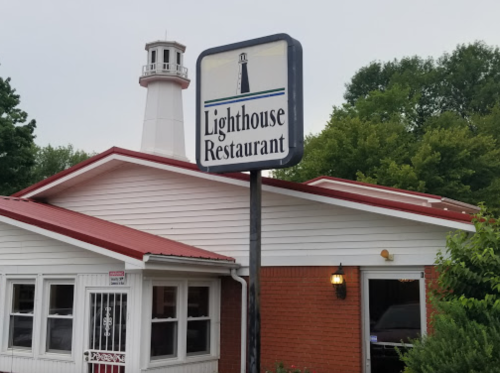 Sign for Lighthouse Restaurant in front of a building with a lighthouse structure and green trees in the background.