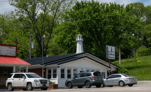 A small restaurant with a lighthouse sign, surrounded by trees and parked cars on a sunny day.