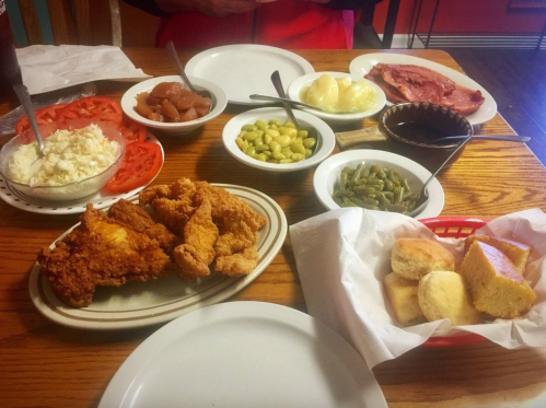 A table spread with fried chicken, sides of vegetables, rice, biscuits, and sliced tomatoes, ready for a meal.