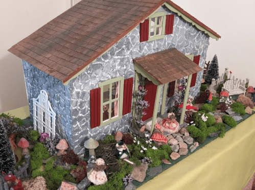 A whimsical miniature house surrounded by colorful mushrooms, greenery, and tiny figurines in a charming garden setting.