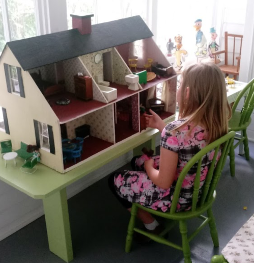 A child plays with a dollhouse on a green table, surrounded by toys in a bright room.