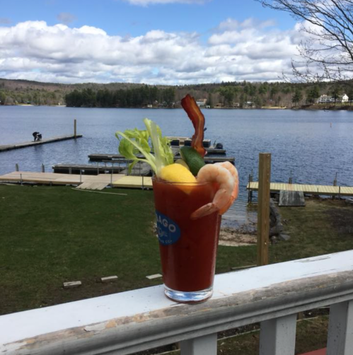A vibrant Bloody Mary garnished with shrimp, lemon, and celery, set against a scenic lakeside view.
