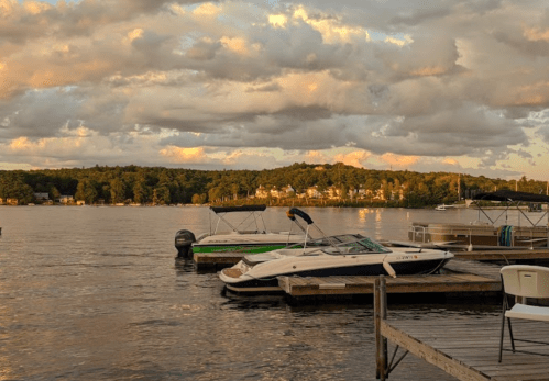 Boats docked at a serene lake during sunset, with clouds reflecting on the water and a wooded shoreline in the background.