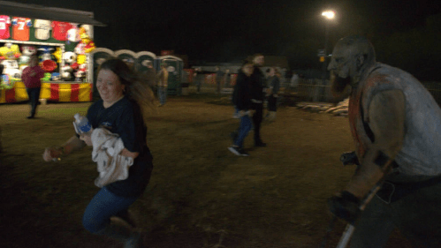 A woman runs away in fear from a costumed figure at a nighttime fair, with games and people in the background.