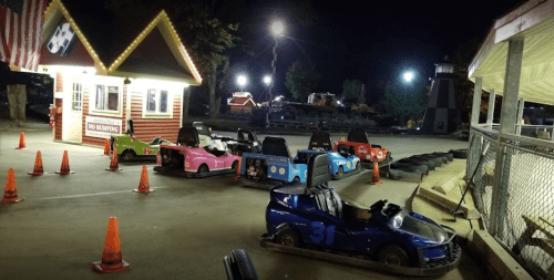 Go-karts lined up at night near a ticket booth, with orange cones and amusement park lights in the background.