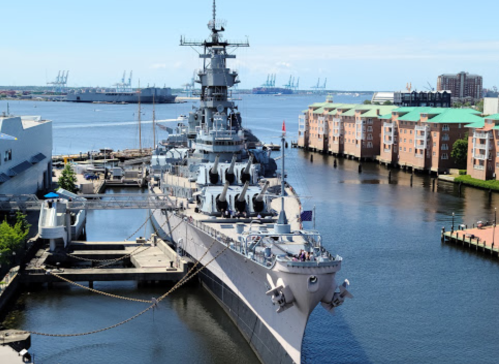 A battleship docked in a harbor, surrounded by buildings and cranes under a clear blue sky.