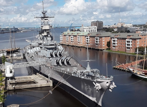 A large battleship docked in a harbor, surrounded by buildings and boats under a partly cloudy sky.