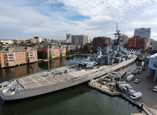 A battleship docked in a city harbor, surrounded by buildings and a calm waterway under a cloudy sky.