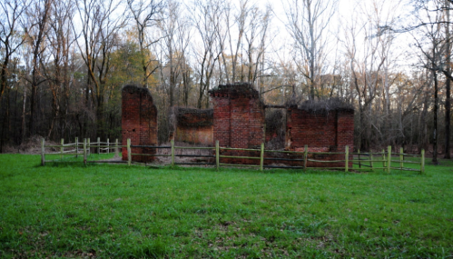 Ruins of a brick structure surrounded by trees and a grassy area, enclosed by a wooden fence.