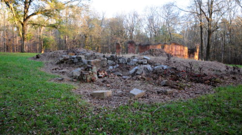 Ruins of a stone structure surrounded by grass and trees, with remnants of brick walls in the background.