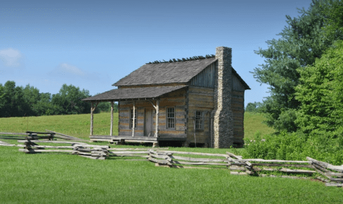 A rustic wooden cabin with a stone chimney, surrounded by green fields and a split-rail fence under a clear blue sky.