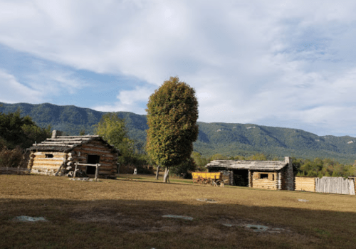 Two rustic log cabins sit in a grassy area with mountains in the background under a partly cloudy sky.