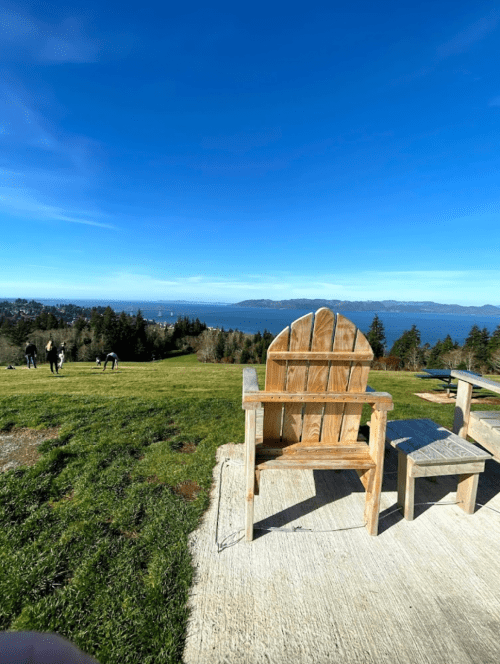 A wooden chair and small table overlook a scenic view of water and distant mountains under a clear blue sky.