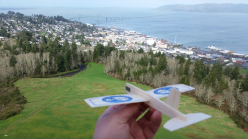 A hand holds a small wooden airplane over a scenic view of a coastal town and water.