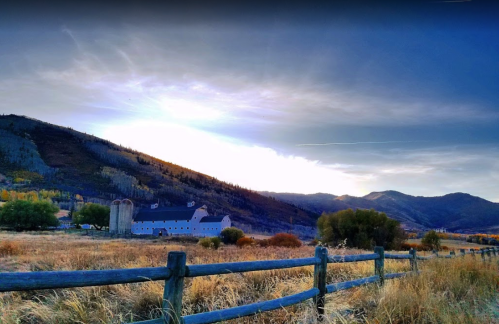 A serene landscape featuring a barn, mountains, and a wooden fence under a cloudy sky at sunset.